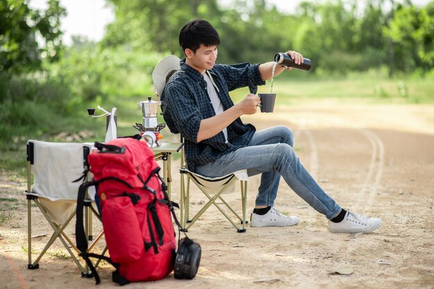 Allegro giovane zaino in spalla uomo seduto davanti alla tenda nella foresta con set da caffè e fare un macinino da caffè fresco durante il viaggio in campeggio durante le vacanze estive