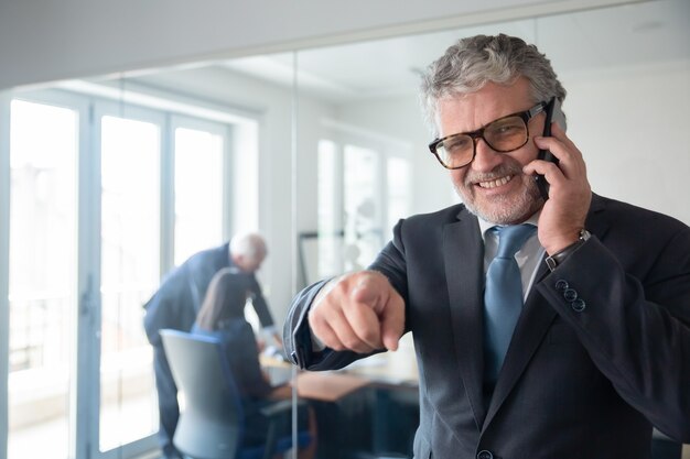 Allegro imprenditore maturo guardando e indicando la fotocamera mentre si è in piedi dalla parete di vetro dell'ufficio, parlando al cellulare e sorridente. Copia spazio. Comunicazione o concetto di lavoro