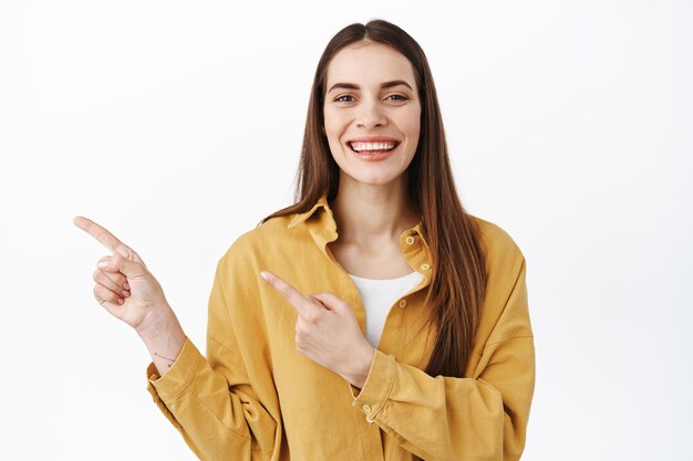 Allegro modello femminile caucasico sorridente e puntando le dita da parte, invitando a visitare il sito Web del negozio, mostrando pubblicità sullo spazio della copia, visualizzando informazioni, in piedi su sfondo bianco