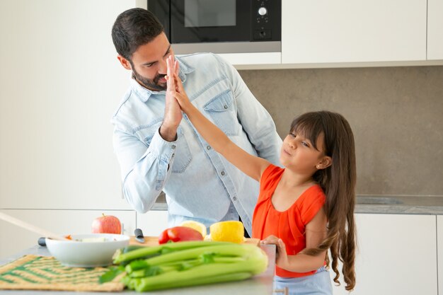 Allegro papà e figlia che danno il cinque mentre cucinano insieme in cucina. Ragazza e suo padre che tagliano le verdure al bancone della cucina. Concetto di cucina familiare