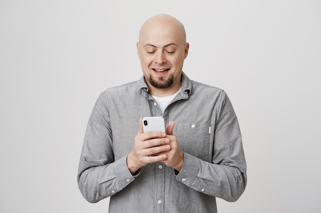 Allegro sorridente uomo adulto calvo messaggistica, utilizzando lo smartphone