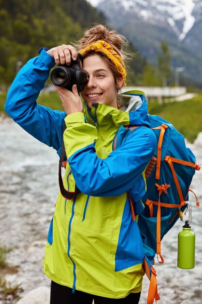 Allegro turista posa sulla vista panoramica, trasporta un grande zaino, scatta foto con la fotocamera, scatta foto del ruscello, indossa una giacca a vento
