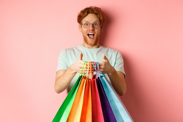 Allegro uomo rosso acquisto di regali, tenendo le borse della spesa e sorridente, in piedi su sfondo rosa.