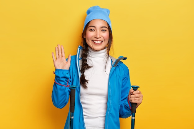 Allegro viaggiatore femminile Asain detiene attrezzatura da trekking, palme onde, saluta amico in montagna, essendo escursionista attivo, sorride piacevolmente, isolato sopra il muro giallo