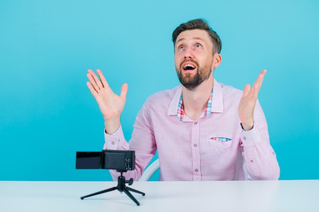 Alzando lo sguardo l'uomo del blogger sta aprendo le mani di hsi sedendosi davanti alla sua mini telecamera su sfondo blu