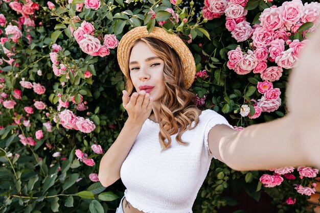 Amabile ragazza bianca che fa selfie sulla natura. Debonair donna riccia che invia un bacio d'aria mentre posa accanto a cespuglio di rose