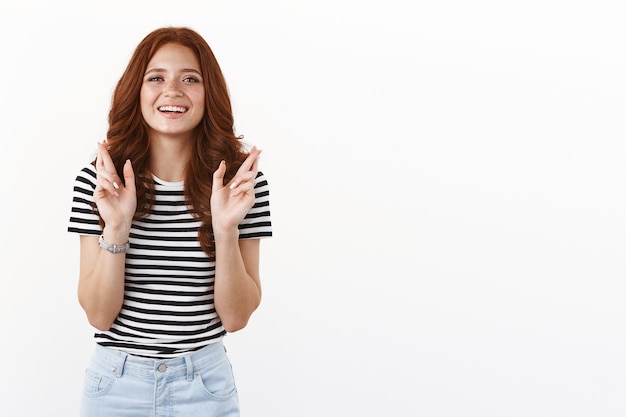 Ambiziosa donna rossa sognante carina in maglietta a righe che ha fede, si spera guardi la macchina fotografica, sorridente anticipando il desiderio che si avvera, incrociando le dita buona fortuna, pregando fortuna, muro bianco