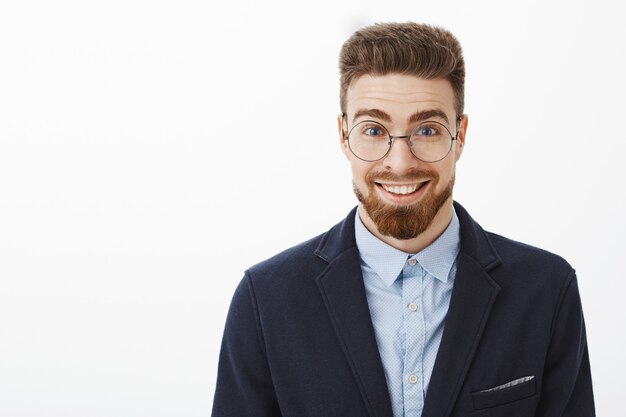 Ambizioso giovane uomo d'affari alla moda intelligente e creativo in occhiali rotondi con barba e occhi azzurri in piedi in abito alla moda che sorride ampiamente sentendosi stupito ed entusiasta di iniziare un nuovo progetto