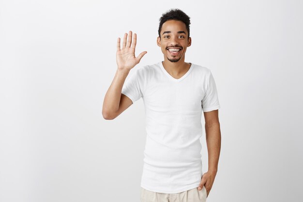 Amichevole bel ragazzo dalla carnagione scura agitando la mano, salutando, salutando o accogliendo persona, sorridente allegro