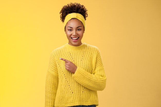 Amichevole bella ragazza entusiasta nera affascinante in maglione con fascia sorridente ampiamente divertito che indica a sinistra un posto fantastico che ti mostra una pubblicità perfetta dello spazio vuoto, in posa su sfondo giallo.