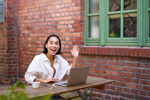 Amichevole donna asiatica seduta con il computer portatile che ti saluta dicendo ciao ciao gesto che ti saluta mentre wo