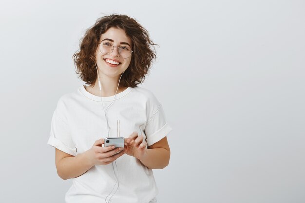 Amichevole donna sorridente in bicchieri tenendo il telefono cellulare, ascoltando musica in auricolari