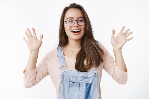 Amichevole e gioiosa giovane donna bruna carina in occhiali e salopette di jeans alzando entrambi i palmi salutando e salutando, salutando facendo un gesto di saluto e sorridendo ampiamente a ogni ospite sul muro grigio.