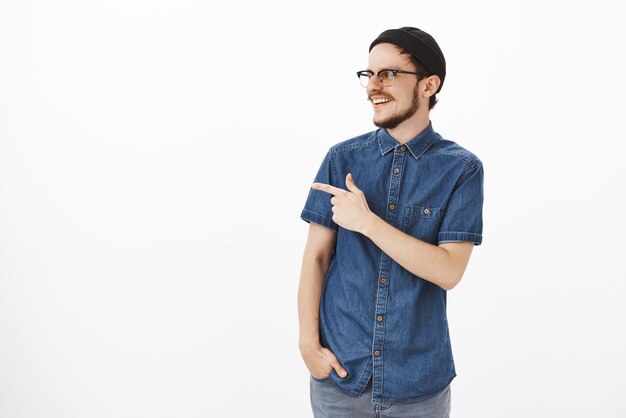 Amichevole e gioioso bel ragazzo con la barba in occhiali e berretto nero hipster che si gira e indica spensierato guardando un ragazzo che fa fantastici trucchi sullo skateboard