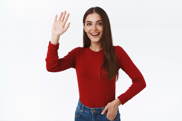 Amichevole ed entusiasta studentessa bruna in maglione rosso che alza la mano in alto cinque o agitando in segno di saluto saluta o saluta con un bel sorriso ampio in piedi su sfondo bianco