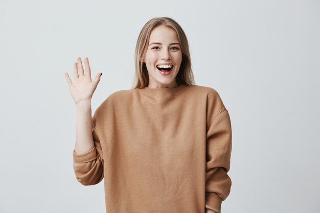 Amichevole femmina bionda positiva che sorride ampiamente e felicemente, saluta con la mano, lieta di incontrarli. Emozioni, sentimenti ed espressione del viso positivi.