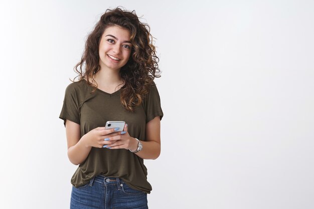 Amichevole gioiosa giovane donna che testa la nuova app per smartphone sorridendo allegramente guarda la fotocamera felice distratta dal raddrizzamento post blog internet, in piedi sfondo bianco, scegli il nuovo negozio online di borse