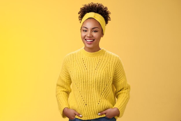 Amichevole in uscita rilassata giovane afro-americana bella ragazza fronte fascia in maglione tenere le mani tasche sorridendo ampiamente divertirsi parlando comunicazione interessante, in piedi muro giallo.