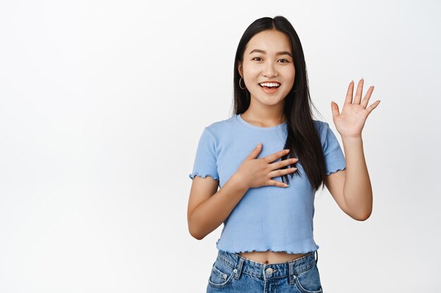 Amichevole ragazza asiatica sorridente che alza la mano pur braccio sul cuore facendo promessa si presenta in piedi in maglietta su sfondo bianco
