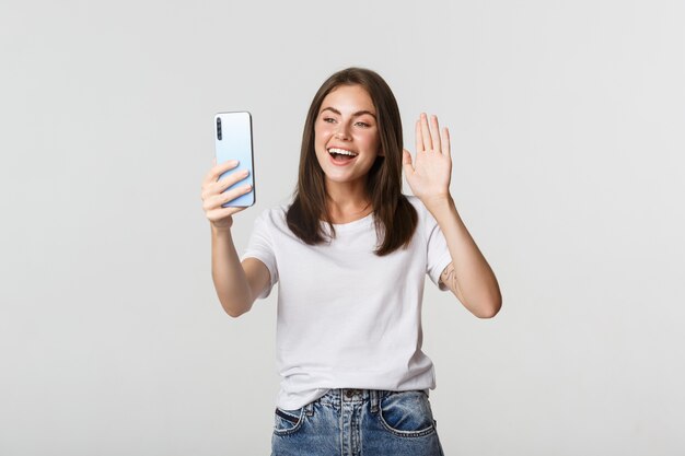 Amichevole ragazza attraente dicendo Ciao, agitando la mano sullo smartphone durante la videochiamata, avendo conversazione.