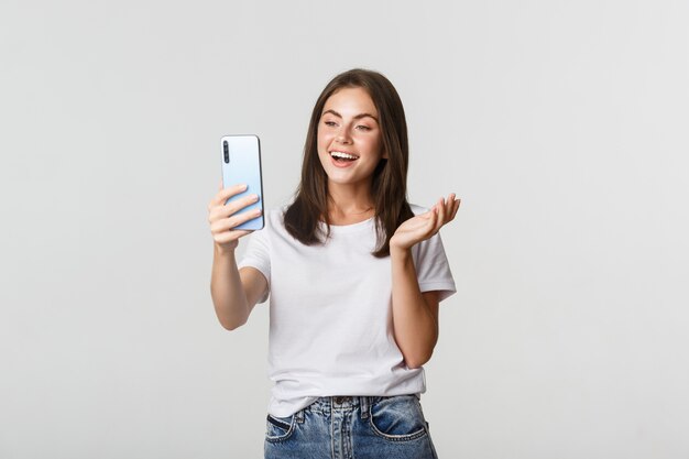 Amichevole ragazza attraente videochiamata amico, sorridente e conversazione, che tiene smartphone, bianco.