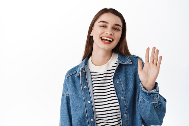 Amichevole ragazza felice agita la mano e saluta, saluta. Donna sorridente che saluta per fare un gesto di saluto, accogliere gli ospiti, felice di vedere qualcuno, in piedi su sfondo bianco