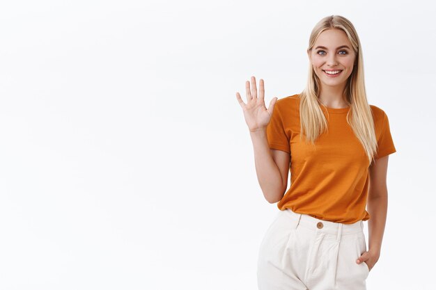 Amichevole, spensierata sfacciata donna bionda attraente in maglietta arancione, pantaloni alzano il palmo e agitando la mano in gesto di saluto, sorridendo con gioia saluta o saluta, benvenuto o ospite, sfondo bianco