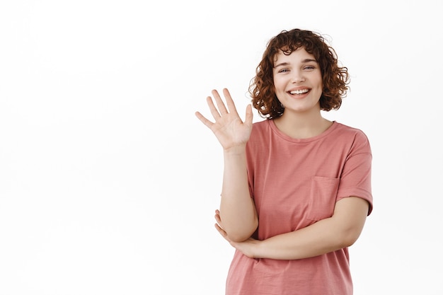 Amichevole studentessa agitando la mano e sorridendo, salutandoti, salutando, accogliendo qualcuno con un gesto ciao, in piedi sul bianco