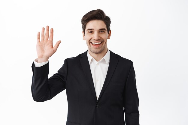 Amichevole uomo di successo sorridente in abito nero, salutando con la mano gesto di saluto, presentandosi, dicendo ciao, benvenuto e salutare qualcuno, sfondo bianco
