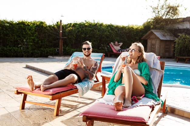 Amici che sorridono, bevendo cocktail, sdraiati su sedie a sdraio vicino alla piscina