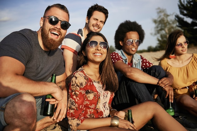 Amici felici seduti con bottiglie di birra sulla spiaggia al tramonto