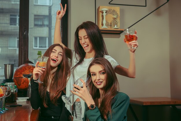 Amici femminili che hanno un drink al bar. Sono seduti a un tavolo di legno con cocktail. Indossano abiti casual.