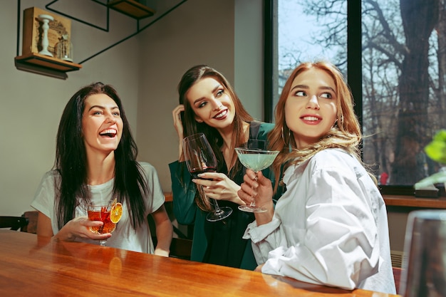 Amici femminili che hanno un drink al bar. Sono seduti a un tavolo di legno con cocktail. Indossano abiti casual.