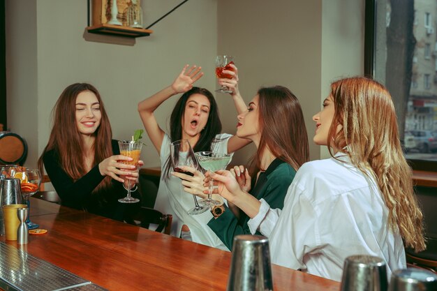 Amici femminili che hanno un drink al bar. Sono seduti a un tavolo di legno con cocktail. Sono bicchieri tintinnanti