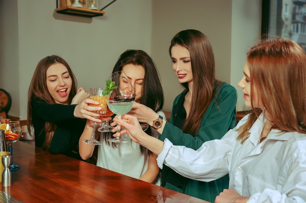 Amici femminili che hanno un drink al bar. Sono seduti a un tavolo di legno con cocktail. Sono bicchieri tintinnanti