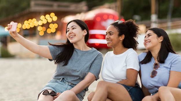 Amici femminili di smiley che si siedono sulla spiaggia e che prendono selfie