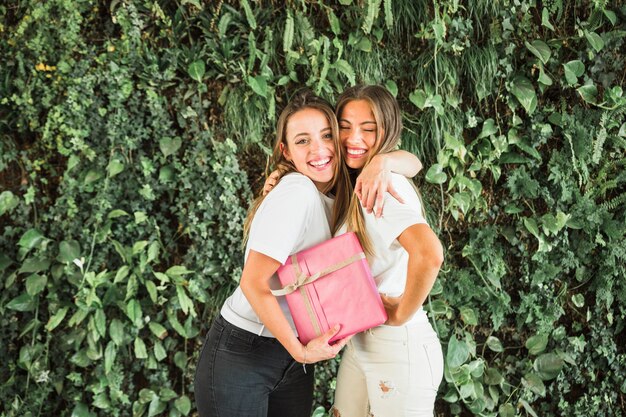 Amici femminili felici con il contenitore di regalo rosa che sta davanti alle foglie verdi