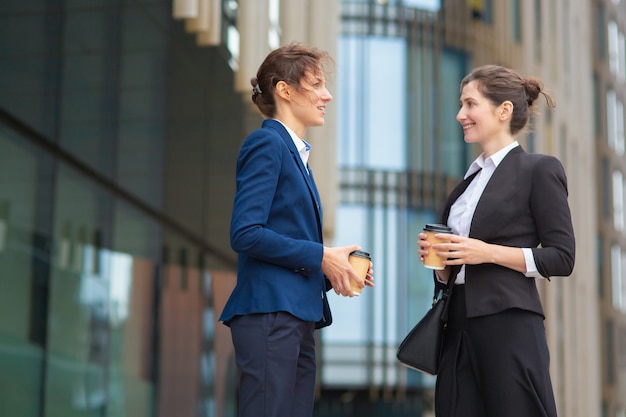 Amici femminili felici dell'ufficio con tazze di caffè da asporto che si incontrano all'aperto, parlano, discutono del progetto o chiacchierano. Vista laterale. Concetto di pausa di lavoro