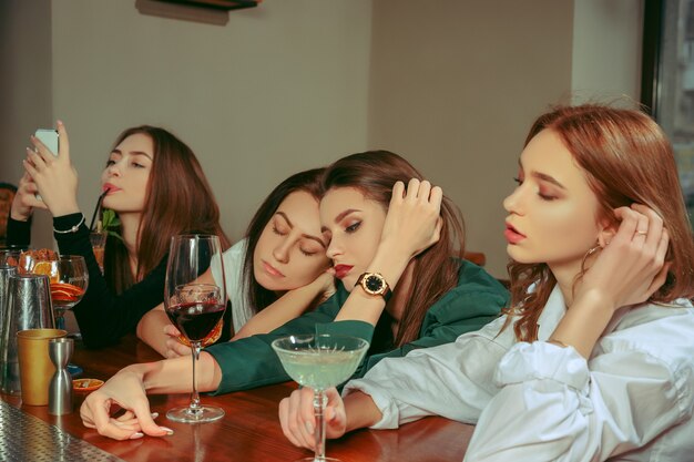 Amici femminili tristi e stanchi che bevono un drink al bar. Sono seduti a un tavolo di legno con cocktail. Indossano abiti casual.