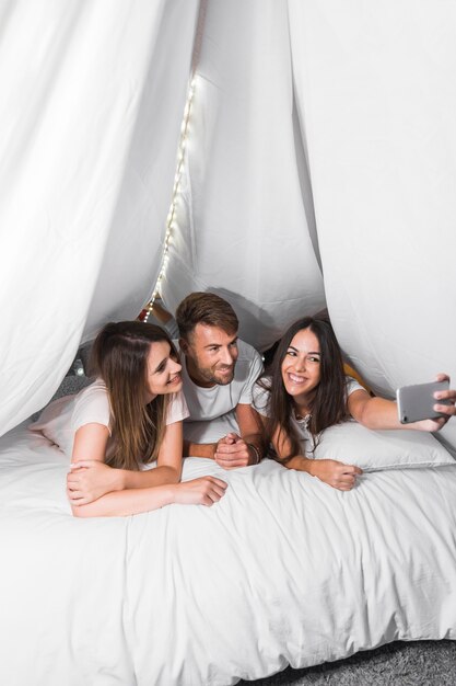 Amici sorridenti che si trovano sul letto che prende selfie dal cellulare
