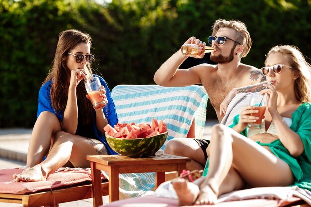 Amici sorridenti, mangiando anguria, bevendo cocktail, rilassandosi vicino alla piscina