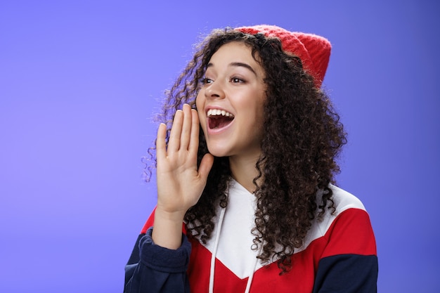 Amico che grida per strada per attirare l'attenzione. Ritratto di donna carina felice spensierata in berretto e felpa che tiene il palmo vicino alla bocca aperta guardando con un sorriso a sinistra come compagno di chiamata con un forte urlo.