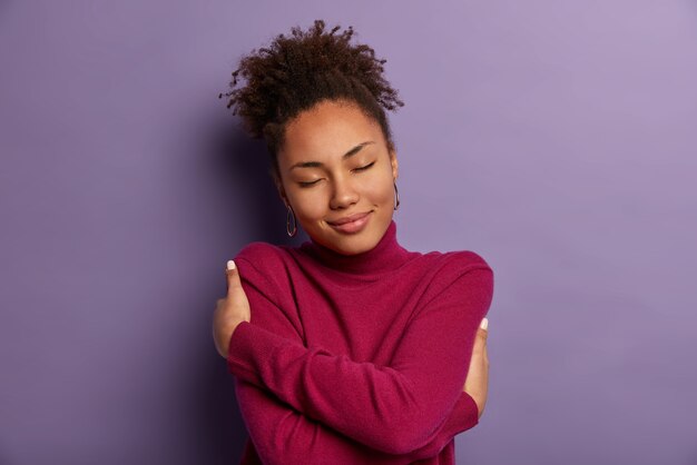 Amo me stessa. Tenera donna romantica abbraccia il proprio corpo, abbraccia se stessa, chiude gli occhi dal piacere, indossa un dolcevita morbido solo per il freddo, si sente a suo agio, sta al chiuso contro il muro viola