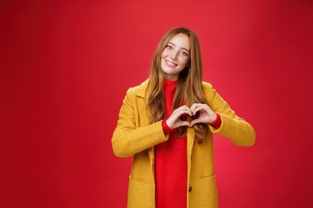 Amore, romanticismo e concetto di caduta. Ritratto di affascinante giovane donna rossa tenera e gentile in cappotto giallo che mostra il gesto del cuore facendo confessione in simpatia, sorridente carino alla telecamera sul muro rosso.