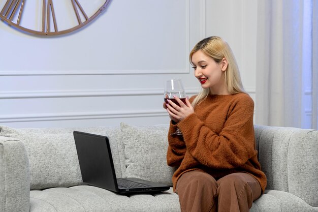 Amore virtuale carina giovane ragazza bionda in un maglione accogliente su un computer a distanza che tiene un bicchiere di vino