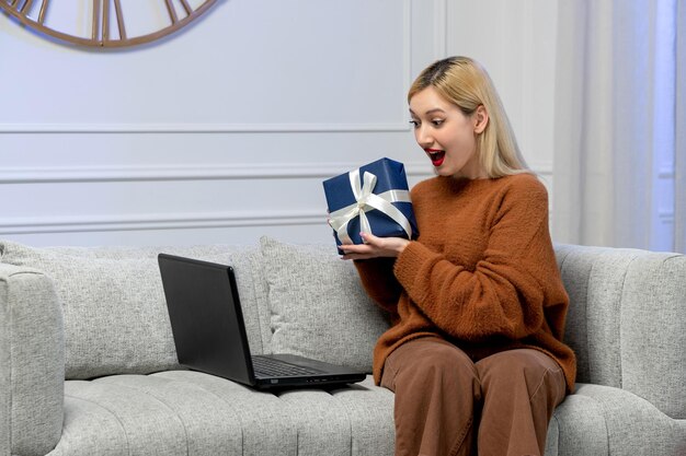 Amore virtuale carina giovane ragazza bionda in un maglione accogliente sulla data del computer a distanza che tiene un regalo blu