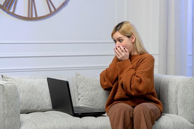 Amore virtuale ragazza bionda carina in maglione accogliente sulla data del computer a distanza che copre la bocca