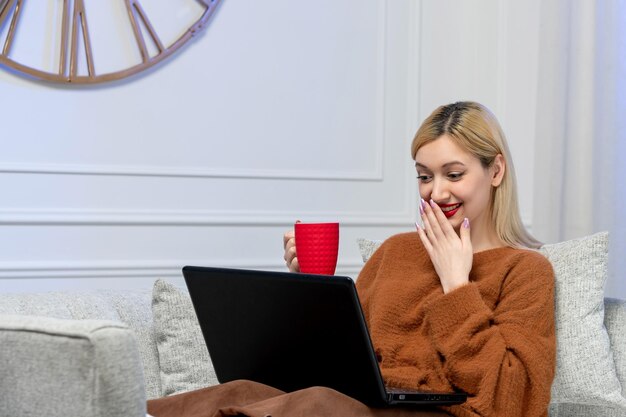 Amore virtuale ragazza bionda carina in maglione accogliente sulla data di distanza del computer che tiene una tazza di tè rossa