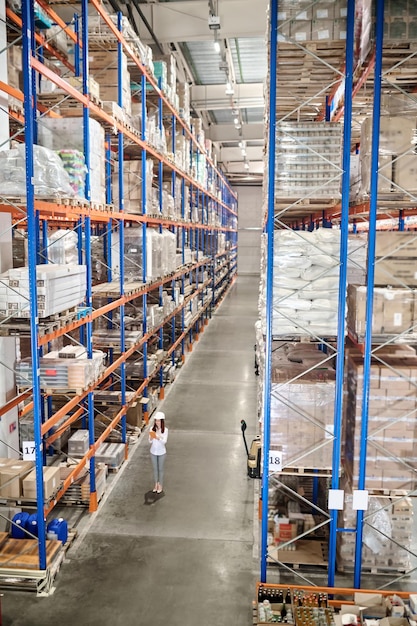 Ampio magazzino. Donna in casco protettivo bianco con tablet in piedi in un grande magazzino industriale nel corridoio tra scaffali alti per merci