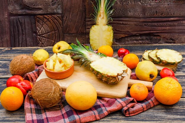 Ananas succosi affettati con noci di cocco, pesche, mele cotogne e agrumi in assi di legno e ciotola sulla superficie di legno di lerciume, panno di picnic e mattonelle di pietra, disposizione piana.